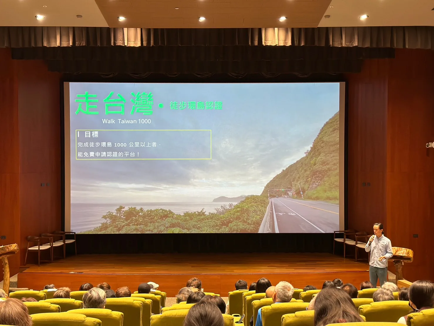 清溪文薈館的大銀幕上是「走台灣・徒步環島認證 Walk Taiwan 1000」簡報,寫著完成徒步環島 1000 公里以上者可免費申請認證,講者站在舞台右側,觀眾背對鏡頭